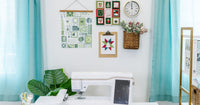 An embroidery machine beneath framed quilt blocks and holiday wall decor, flanked by teal curtains in a bright sewing room.