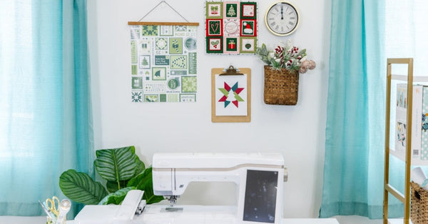 An embroidery machine beneath framed quilt blocks and holiday wall decor, flanked by teal curtains in a bright sewing room.