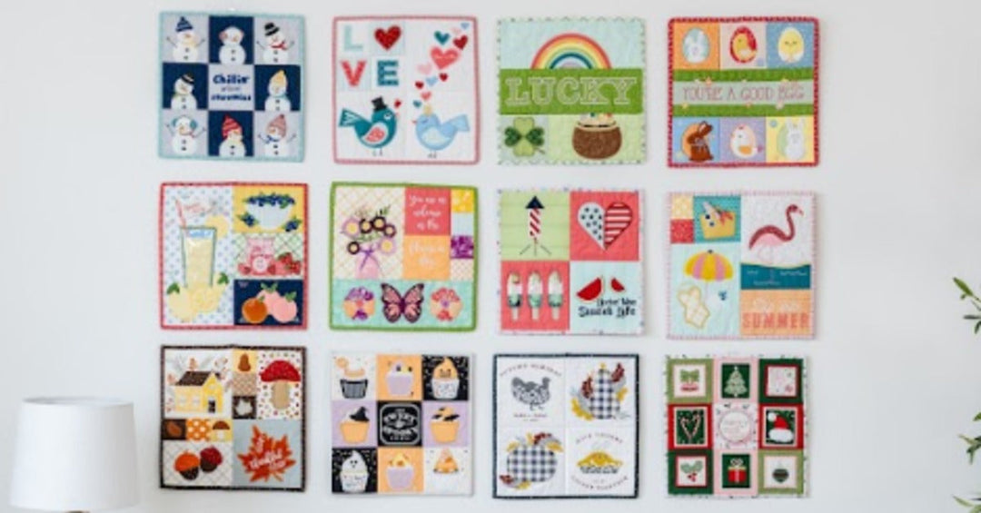 A bright living room with a white sofa beneath a grid of colorful embroidered quilt blocks displayed on a large wall.