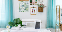 A white embroidery machine on a craft table beneath quilt wall art, a clock, and bright turquoise curtains.