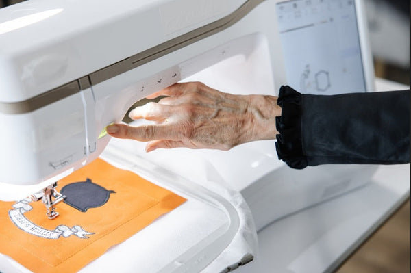Hand adjusts fabric beneath a machine embroidery needle as the machine stitches an orange quilt block.