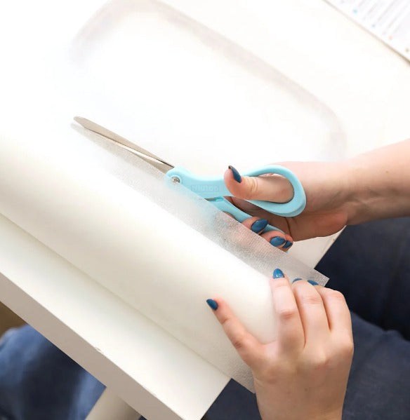 Hands use blue-handled scissors to cut a sheet of white embroidery stabilizer on a table for a machine embroidery project.