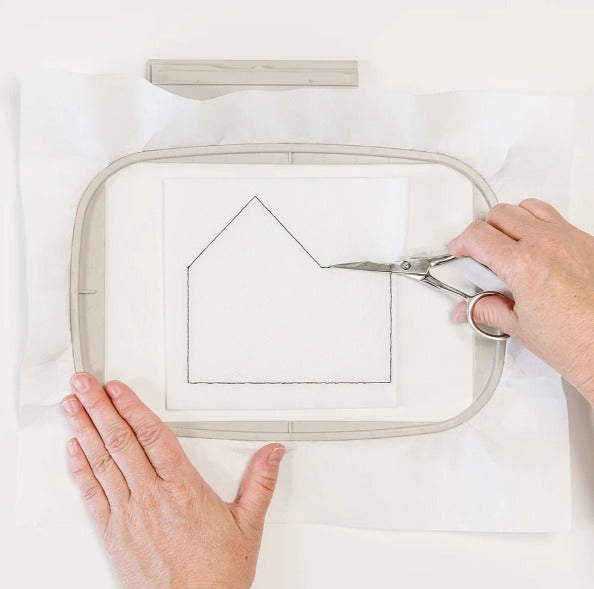 Hands hold an embroidery hoop while scissors trim white stabilizer around a stitched house-shaped design.