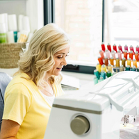 Woman with blonde hair sits at an embroidery machine in a bright sewing room with colorful thread spools behind her.