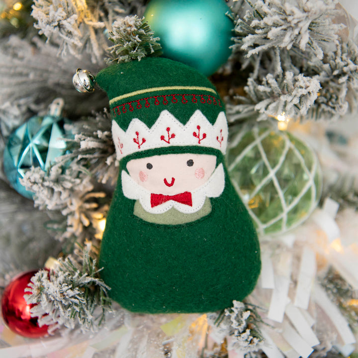 A green felt Christmas elf stuffy with a green and white patterned hat, red bow tie, and embroidered face, hanging on a flocked Christmas tree with colorful ornaments.