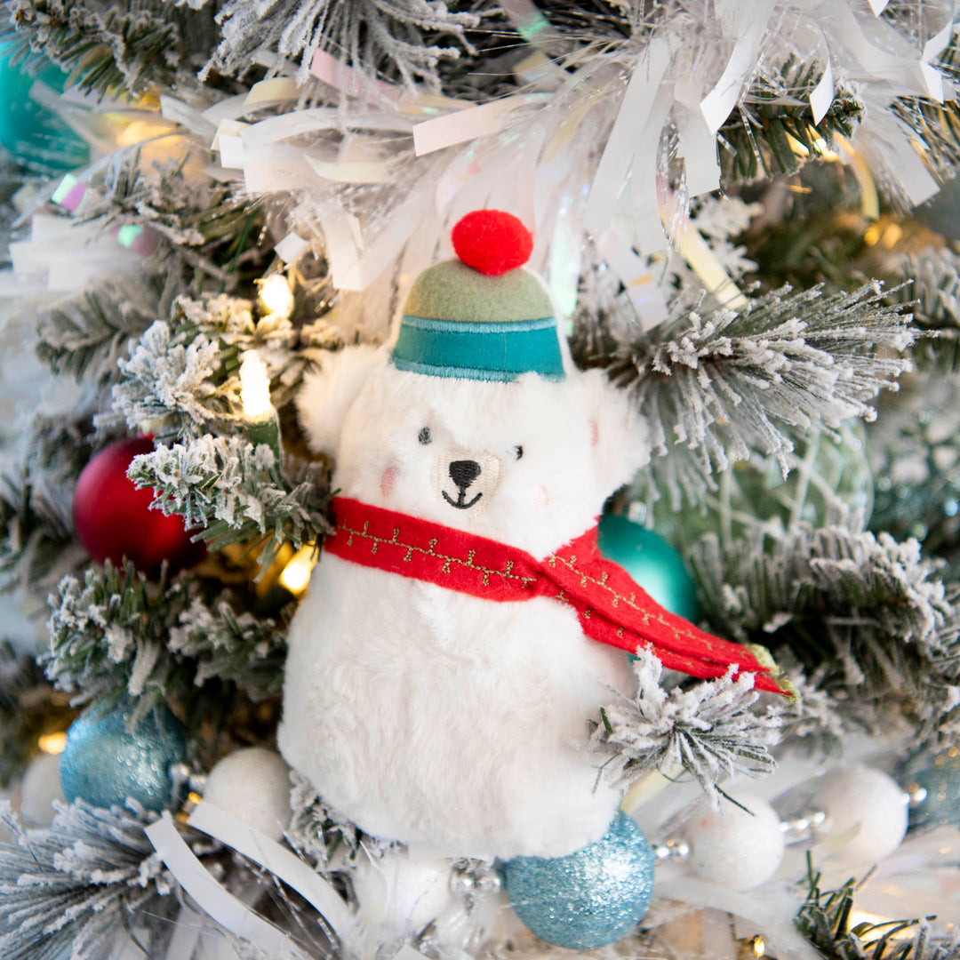 A plush white polar bear stuffy hanging on a flocked Christmas tree, wearing a green and teal hat with a red pom-pom and a red scarf.