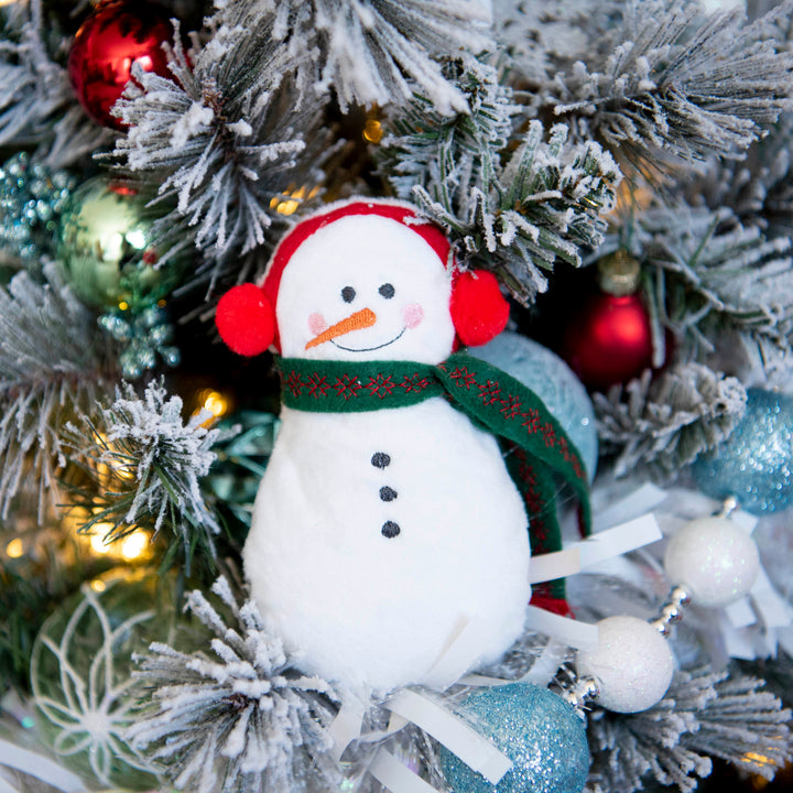 A plush snowman stuffy with red earmuffs and a green patterned scarf, nestled among a flocked Christmas tree branch with colorful ornaments and warm string lights.