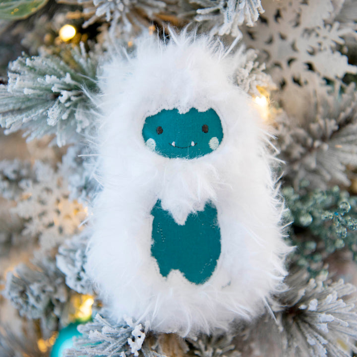 A fluffy white stuffy shaped like a cute cartoon yeti with teal facial features and a teal chest patch, hanging on a frosted Christmas tree with warm lights.