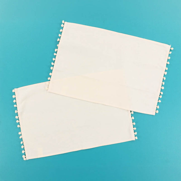 Two ivory fabric placemats with pom-pom edges on a blue background