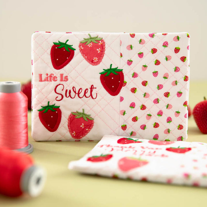 Mug rug with strawberry pattern and 'Life is Sweet' text on a table with spools of thread and strawberries spread about.