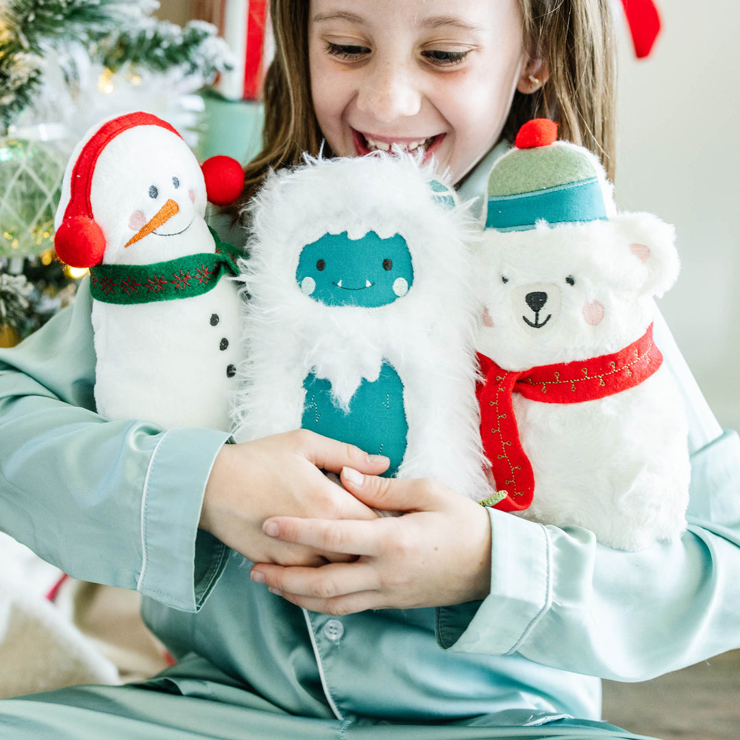A smiling child in light blue pajamas holds three plush Christmas-themed finger puppets: a snowman with a red hat and green scarf, a blue yeti in white fur, and a white polar bear with a red scarf and green hat.