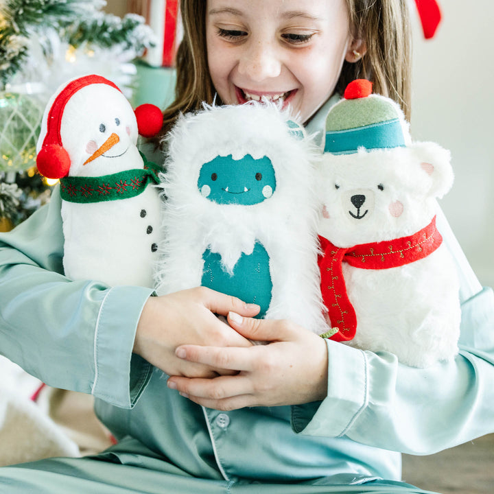 A smiling child in light blue pajamas holds three plush Christmas-themed finger puppets: a snowman with a red hat and green scarf, a blue yeti in white fur, and a white polar bear with a red scarf and green hat.